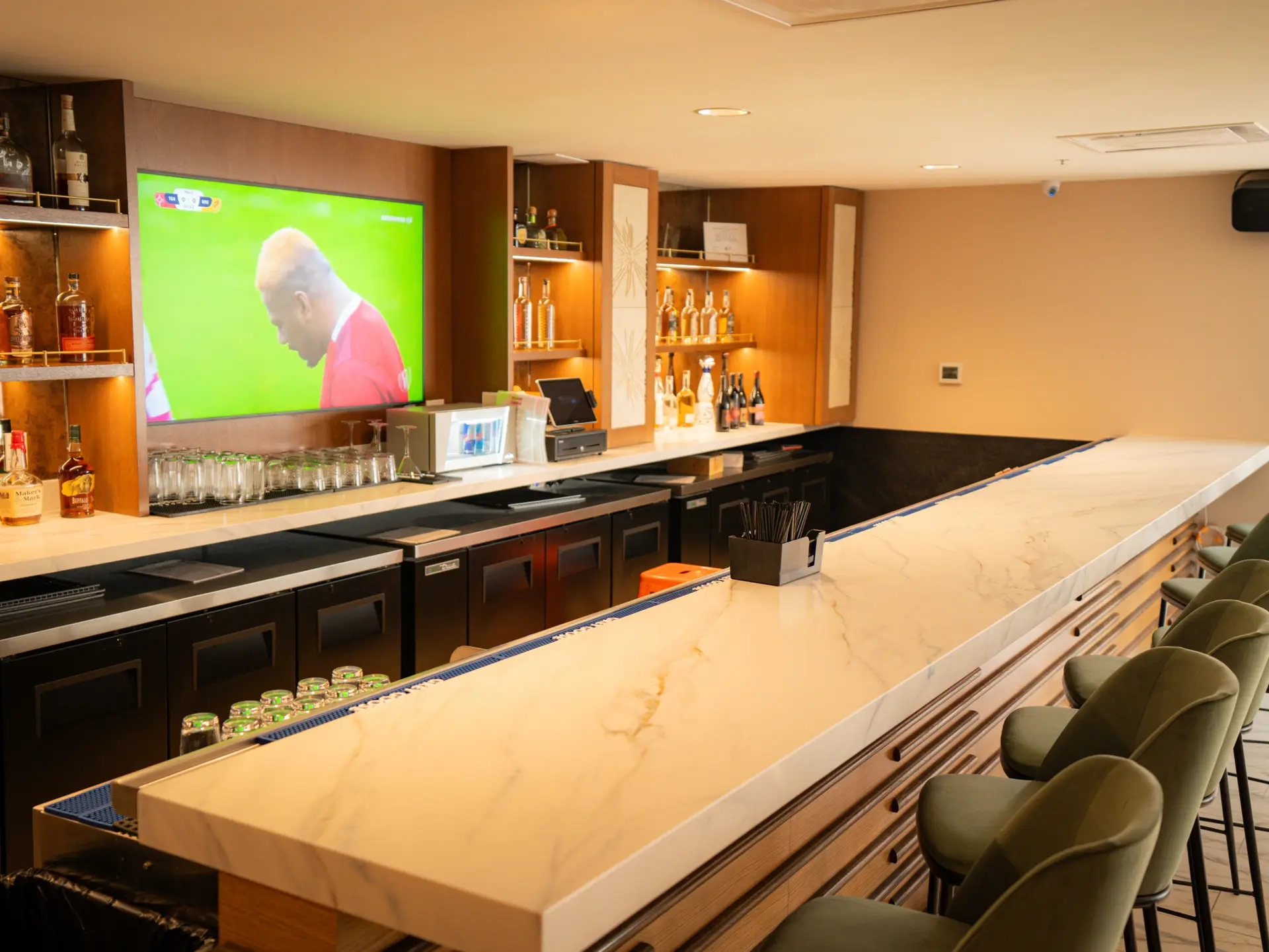 Long white veined stone bar top with green bar stools and lit back-bar shelving