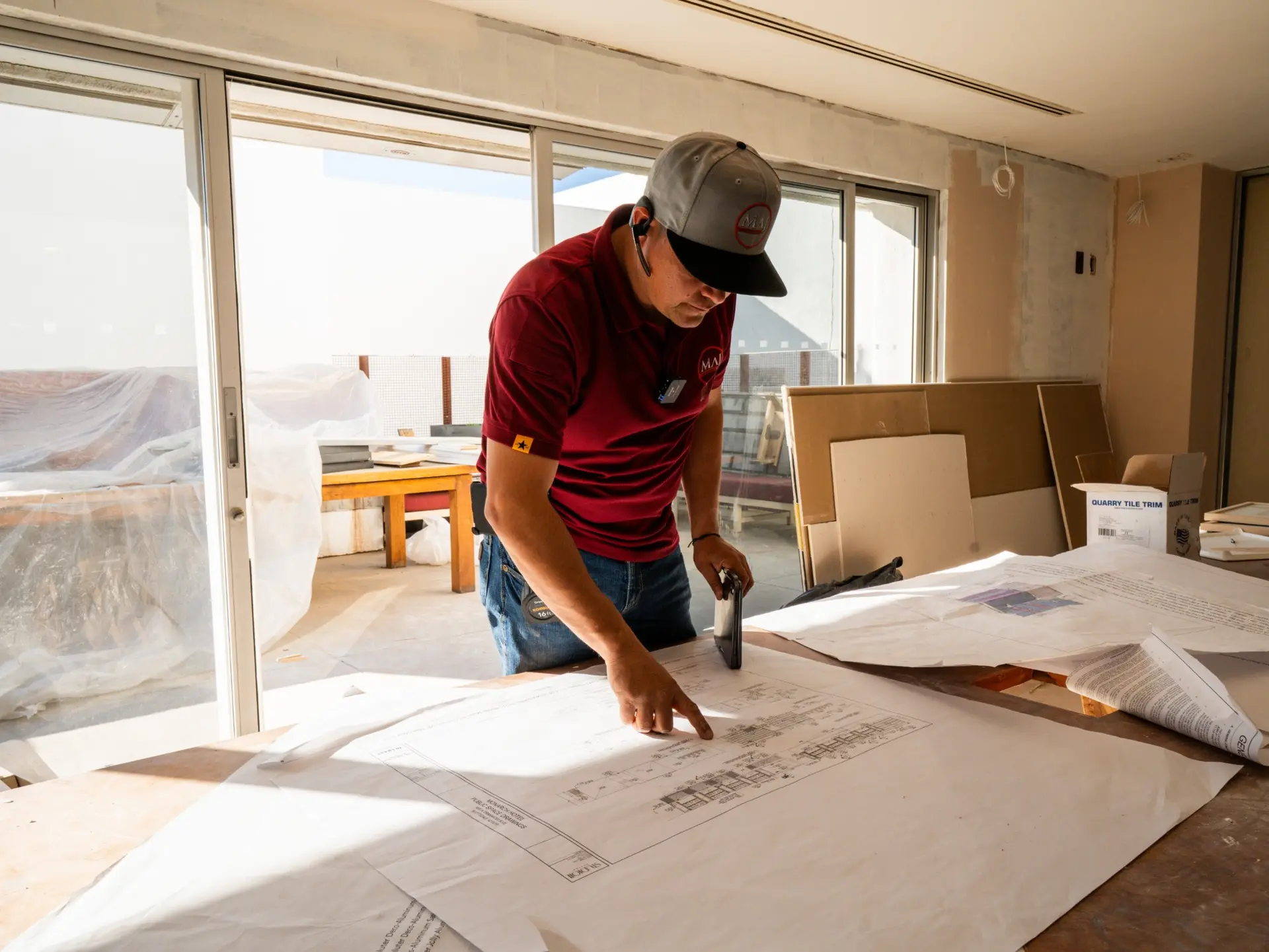 A MAJ Countertops team member in a company shirt and cap reading architectural drawings spread across a table on a job site