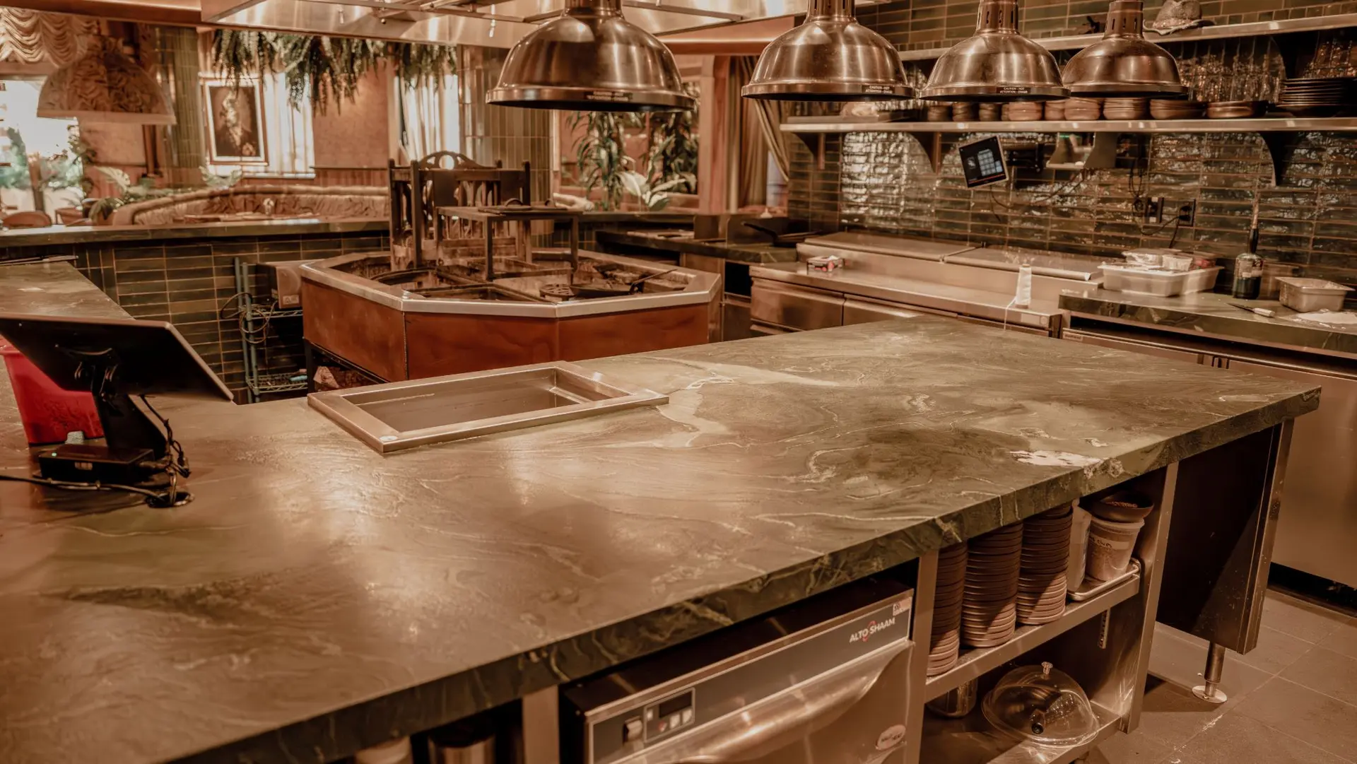 Restaurant kitchen counter in green stone with white veining, a drop-in steel well and pans stacked on open shelves below